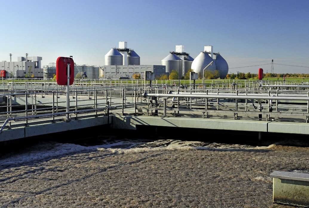 Blick auf ein Belebungsbecken in der Kläranlage mit Faultürmen im Hintergrund.